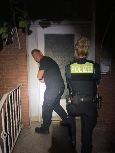 Zwei Polizeibeamte stehen vor einer Tür bei Nacht, einer öffnet die Tür.