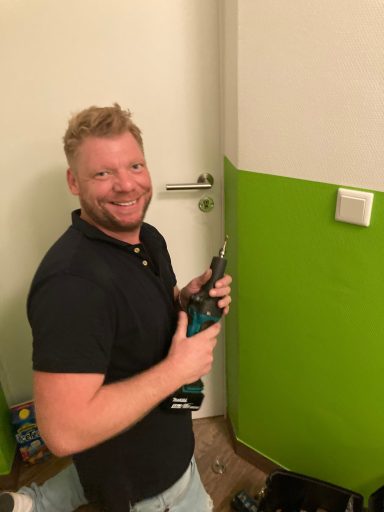 Mann mit Bohrmaschine, der an einer Wand mit grüner Farbe lächelt.